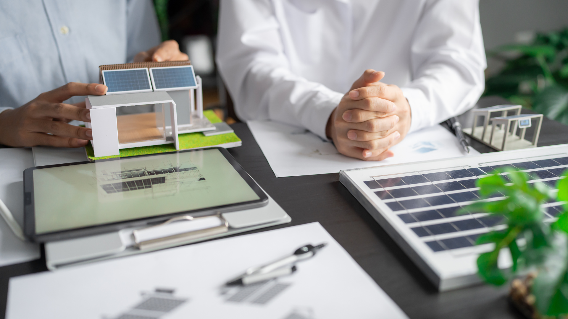 Maquette de bâtiment avec panneaux solaires en toiture, posée sur une table de travail à côté d'une tablette affichant un plan et d'un panneau photovoltaïque